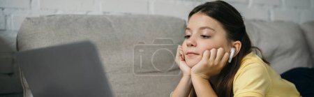 preteen girl in wireless earphone looking at blurred laptop on sofa at home, banner
