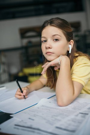Photo for Preteen girl in wireless earphone holding pen and looking at camera while doing homework - Royalty Free Image