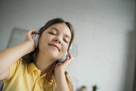 low angle view of pleased preteen girl with closed eyes listening music in headphones