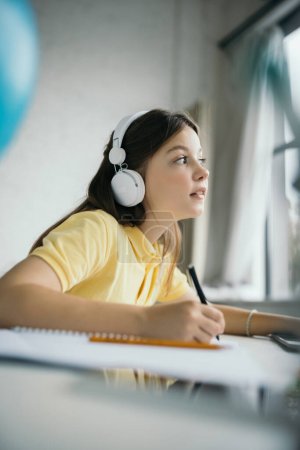 pensive schoolgirl in headphones holding pen and looking away while doing homework