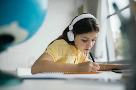 Téléchargez les photos : Écolière en casque écrit avec stylo tout en apprenant à la maison sur le premier plan flou - en image libre de droit