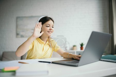 Photo for Smiling girl in headphones waving hand near laptop during online lesson at home - Royalty Free Image
