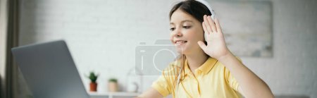 preteen girl in headphones waving hand and smiling during video call on laptop, banner