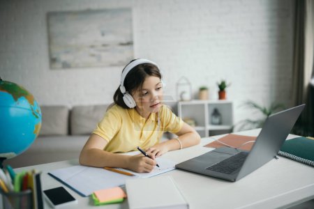 girl in headphones looking at computer and writing in copybook during homeschooling