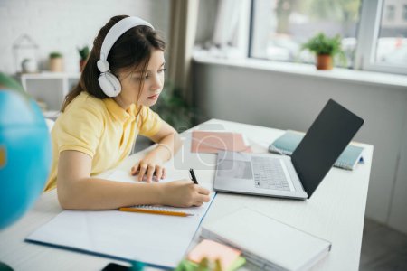 Kind mit Kopfhörer schaut auf Laptop und schreibt zu Hause in Notizbuch