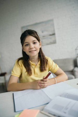 Photo for Cheerful girl holding pens and looking at camera near empty copybook at home - Royalty Free Image