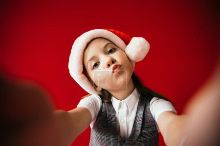 girl in santa hat pouting lips while looking at camera on blurred foreground isolated on red