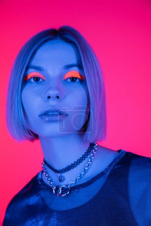 Porträt einer jungen Frau mit Neon-Make-up, die in blaues Licht auf tiefrosa isoliert in die Kamera blickt