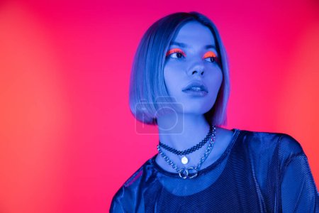 modische Frau mit Neon-Make-up schaut weg in blauem Licht auf korallenrosa Hintergrund