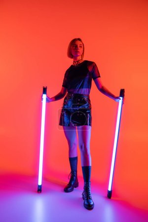 full length of slender and trendy woman holding neon lamps and looking away on coral and purple background