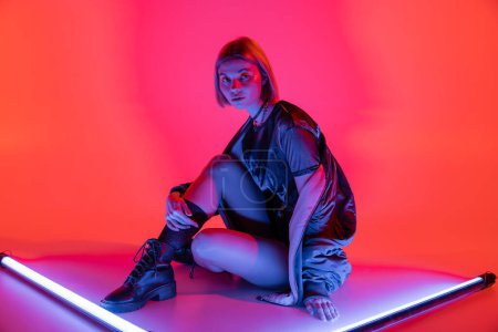 young and fashionable woman sitting near neon lamps and looking at camera on purple and coral background