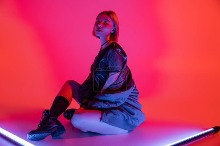 young woman in leather boots sitting near bright neon lamps on purple and coral background
