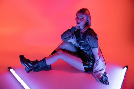 full length of woman in leather boots and mini skirt looking at camera near neon lamps on purple and coral background