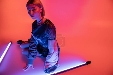Téléchargez les photos : Jeune femme en vêtements élégants regardant la caméra tout en étant assis près de lampes au néon lumineux sur fond violet et corail - en image libre de droit