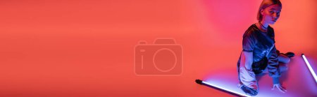 Téléchargez les photos : Femme à la mode assise près de lampes fluorescentes violettes sur fond corail, bannière - en image libre de droit