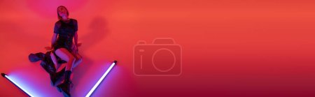 Photo for High angle view of young woman in boots and mini skirt posing near shining neon lamps on purple and red background, banner - Royalty Free Image