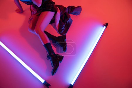 Draufsicht der gestutzten Frau in schwarzen Lederstiefeln in der Nähe von lila Neonlampen auf karminrotem Hintergrund