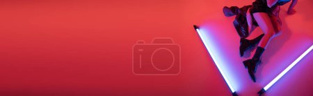 Téléchargez les photos : Vue du dessus de la femme coupée en bottes assis près de lampes au néon violet sur fond rouge carmin, bannière - en image libre de droit