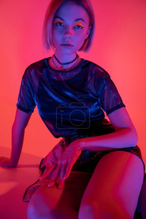 stylish woman with bright neon makeup sitting in purple light on coral red background