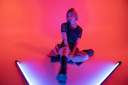 full length of woman in trendy boots sitting near purple neon lamps and looking away on coral background