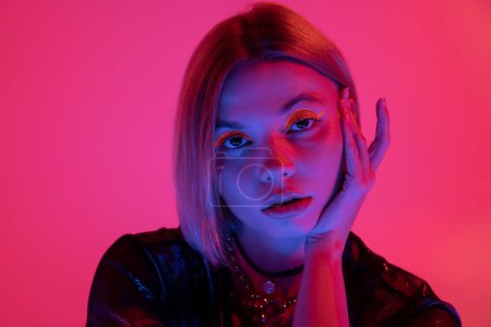portrait of pretty woman with neon makeup holding hand near face in blue light on deep pink background