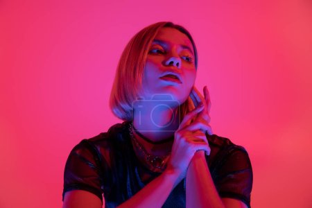 woman in neon light touching face and looking away on deep pink background