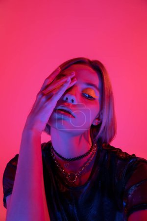 Frau mit Neon-Make-up und Halsketten verschleiert Gesicht mit Hand in lila Licht auf tiefrosa Hintergrund