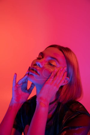 sensual woman with bright neon makeup touching face while posing with closed eyes on coral pink background 