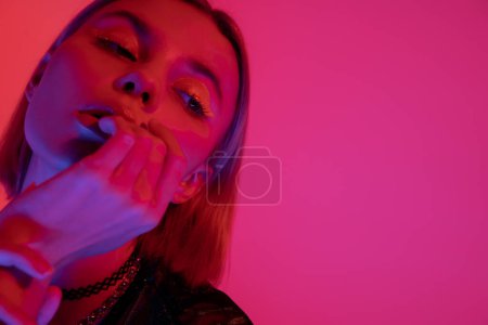 Foto de Retrato de mujer joven con maquillaje de neón sosteniendo la mano cerca de la cara sobre fondo rosa profundo - Imagen libre de derechos