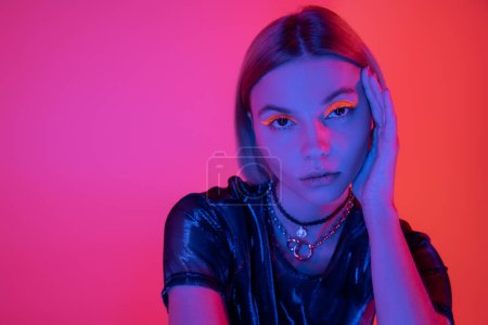 portrait of stylish woman with bright neon makeup and necklaces in blue light on deep pink and coral background