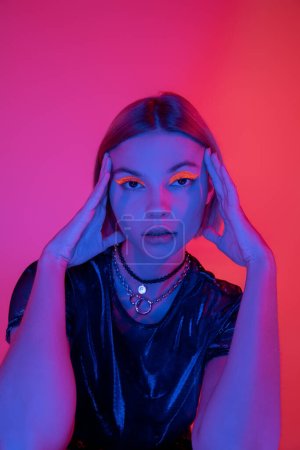 Trendfrau in hellem Neon-Make-up und Halsketten posiert mit händennahem Gesicht auf dunkelrosa und korallenfarbenem Hintergrund
