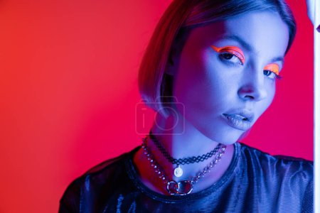 Frau in Halsketten und grellem Neon-Make-up blickt in die Kamera in blauem Licht auf korallenrosa Hintergrund