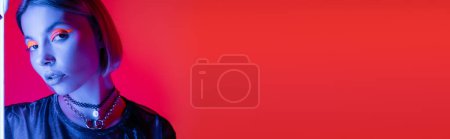 woman with neon makeup looking at camera in blue light on coral red background, banner 