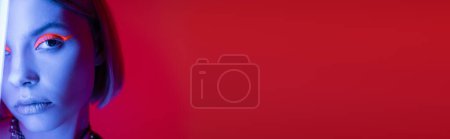 Photo for Partial view of young woman in blue neon light looking at camera on carmine red background, banner - Royalty Free Image