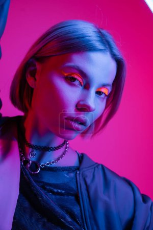 stylish woman in necklaces and glowing neon makeup posing in blue light on deep pink background