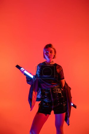 trendy woman in leather mini skirt standing with neon lamp and looking at camera isolated on coral red