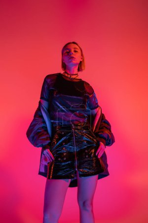 low angle view of woman in leather mini skirt looking at camera in neon light on coral and pink background 