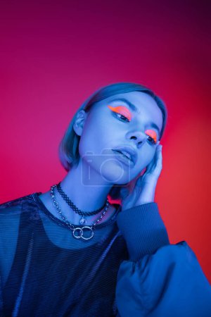 trendy woman in necklaces and neon makeup holding hand near face on pink and red background
