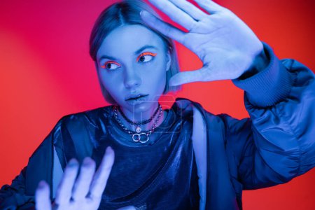 young woman in neon makeup looking away while posing with outstretched hands on  coral red background