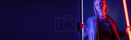 stylish woman with shining neon lamps looking at camera on dark blue background, banner
