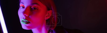 portrait of young woman with neon makeup posing in purple light of neon lamp on black background, banner