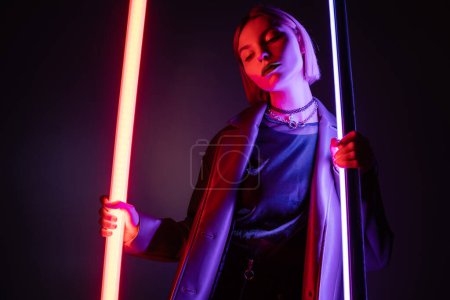 junge und stilvolle Frau posiert mit hellen Neonlampen auf dunkelviolettem Hintergrund