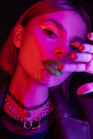 portrait of young woman with bright neon makeup holding hand near face in purple light on black background