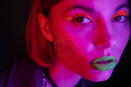 Photo for Close up portrait of woman with glowing neon makeup in purple light on black background - Royalty Free Image