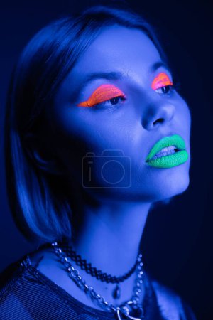 portrait of stylish woman in colorful neon makeup and necklaces looking away on dark blue background