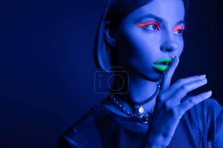 portrait of woman in neon makeup touching green lips while showing hush sign on dark blue background