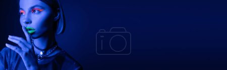 Photo for Young woman with bright neon visage touching green lips and showing hush gesture on dark blue background, banner - Royalty Free Image