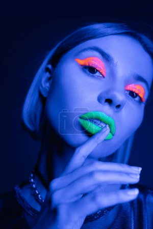 portrait of young woman with colorful neon makeup touching green lips isolated on dark blue
