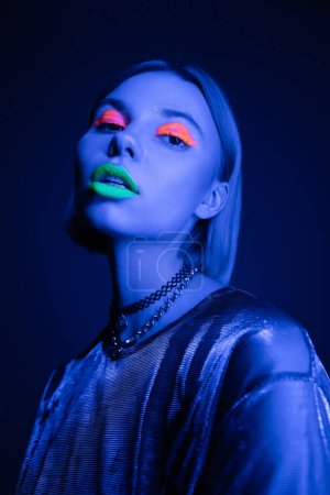 trendige Frau in glänzender Bluse und leuchtendem Neon-Make-up mit Blick in die Kamera auf dunkelblauem Hintergrund