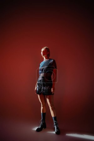 full length view of woman in leather boots and mini skirt standing in sunlight on burgundy background